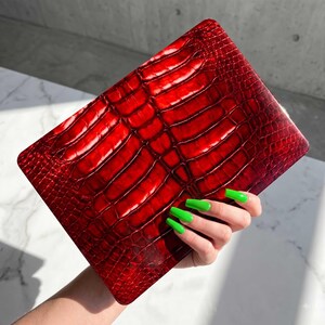 May include: A rectangular clutch bag featuring a glossy, deep red finish with a textured, embossed pattern that mimics the appearance of reptile scales. The surface has a reflective quality, showing variations in light and shadow across the scales.