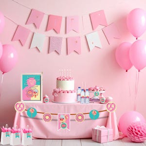 Puede incluir: Una escena de fiesta rosa con una pancarta, globos y una mesa preparada para una celebración. La mesa presenta una tarta, cupcakes y una guirnalda con diseños de donuts y piruletas. La escena incluye bolsas de regalo y un letrero enmarcado.