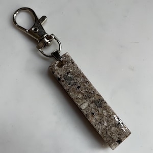 Puede incluir: Un llavero rectangular con una apariencia moteada similar a la piedra en tonos grises, marrones y blancos. Está unido a un cierre de metal plateado. El llavero está diseñado para guardar llaves o sujetarlo a una bolsa.