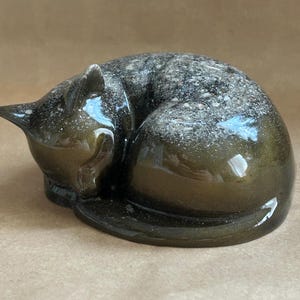 May include: A smooth, dark olive-green resin figurine of a sleeping cat curled into a ball. The surface features a glossy finish with embedded light-coloured, speckled particles concentrated along the back, creating a textured, shimmering appearance.