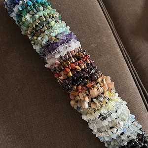 Op de afbeelding: Een verzameling edelsteen chip armbanden in een regenboog van kleuren, waaronder blauw, groen, paars, oranje en wit. De armbanden zijn gestapeld op een zwarte cilinder, die de verscheidenheid aan stenen laat zien.