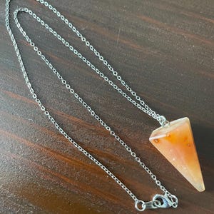 May include: A silver chain necklace with a triangular, orange-colored pendant. The pendant has a pointed end and a translucent appearance. The necklace is displayed on a dark brown wooden surface.