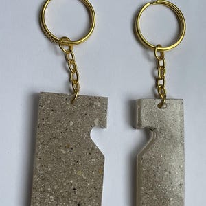 Op de afbeelding: Twee bijpassende sleutelhangers. Elke sleutelhanger heeft een rechthoekige, gespikkelde, grijze hanger met een hartvormige uitsparing. De hangers zijn bevestigd aan goudkleurige sleutelringen met kleine kettingen. De achtergrond is wit.