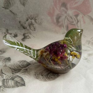 May include: A clear resin bird figurine containing embedded dried flowers and greenery. The interior features a mix of purple, yellow, and white blossoms along with a green fern leaf extending into the tail, all encased in a smooth, transparent form.