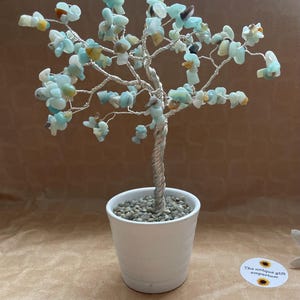 May include: A decorative tree sculpture with a silver wire trunk and branches, adorned with light blue and tan gemstone leaves. The tree sits in a white ceramic pot filled with small pebbles. The Unique Gift Emporium logo is visible.