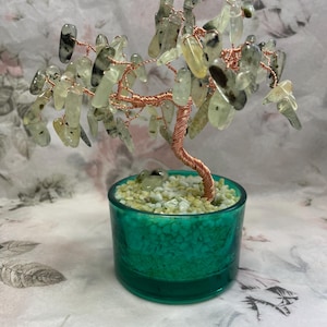 May include: A handcrafted tree sculpture featuring a copper wire frame and light green and black gemstone leaves. The tree is planted in a teal glass pot filled with small stones. The background is a patterned fabric, creating a natural, decorative piece.