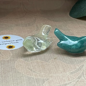 May include: Two small bird figurines. One is clear, the other is teal with a speckled appearance. The birds are positioned on a light-coloured surface. A circular sticker with the text "The unique gift emporium" is also visible.