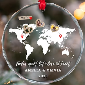 May include: A clear, round glass ornament with a world map design. The ornament features photos in heart-shaped frames, red pin markers, and the text "Miles apart, but close at heart. AMELIA & OLIVIA 2025." A red ribbon is attached for hanging.