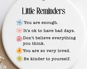 May include: A white ceramic disc ornament with the text "Little Reminders" and inspirational quotes. The ornament has a ribbon for hanging and is decorated with watercolor flower illustrations. The words "FREE GIFT BOX" are in a black box.