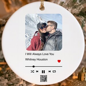 May include: White ceramic Christmas ornament with a photo of a couple kissing. The ornament features the song title "I Will Always Love You" by Whitney Houston, a play bar, and a heart icon. A gold string is attached for hanging.