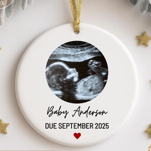 May include: A white ceramic ornament with a gold ribbon for hanging. The ornament features a sonogram image in a circular frame, with the text "Baby Anderson DUE SEPTEMBER 2025" and a red heart. Gold star decorations are scattered around the ornament.