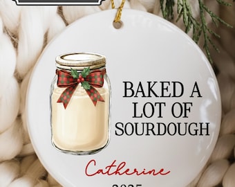 May include: White ceramic ornament with a jar illustration, a red and green bow, and the text "BAKED A LOT OF SOURDOUGH." It also includes the name "Catherine" and the year "2025."