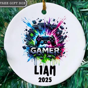 May include: White ceramic ornament with a colorful gamer design. The design features a video game controller, the word "GAMER", the name "LIAM", and the year "2025". The ornament has a gold string for hanging. The text "FREE GIFT BOX" is in the top left corner.