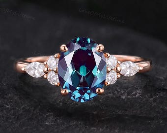 May include: A rose gold ring featuring a large oval-cut color-changing gemstone in the center, flanked by two smaller round-cut stones and two pear-cut stones on each side, set on a thin, polished rose gold band.
