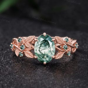Può includere: Un anello in oro rosa con una pietra centrale in agata muschiata dal taglio ovale con inclusioni verdi visibili. La fascia è decorata con motivi a foglia in metallo e piccole pietre verdi rotonde incastonate lungo i lati della montatura.