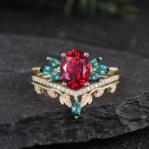 May include: A gold ring set featuring a central oval-cut red gemstone flanked by marquise-cut green stones. The band includes a V-shaped nesting ring adorned with small white stones and a leaf-like metal texture along the lower edge of the band.