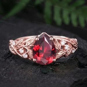 Pode incluir: Um anel de ouro rosa com uma grande pedra preciosa granada vermelha em forma de pera como peça central. A banda é desenhada com motivos de folhas intrincados e acentuada com pequenas pedras brancas redondas e claras incrustadas nas laterais.