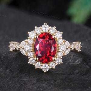Könnte beinhalten: Ein Goldring mit einem zentralen, oval geschliffenen, tiefroten Edelstein, der von einem Kranz aus kleinen runden und marquiseförmigen klaren Steinen umgeben ist. Die Ringschiene besteht aus Gelbgold und ist seitlich mit weiteren klaren Steinen besetzt.