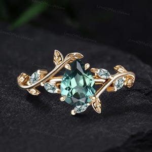 May include: A gold ring featuring a central pear-shaped green gemstone surrounded by a vine-like band design. The band incorporates smaller, marquise-shaped moss-like stones set within gold leaf motifs that extend from the central setting.