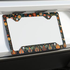 May include: A decorative license plate frame featuring a dark background with a floral and moth pattern. The frame is mounted on the back of a white car. The design includes orange, yellow, and green flowers, and various moth illustrations.
