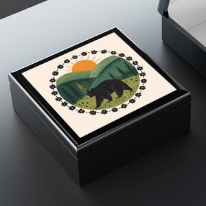 Könnte beinhalten: Zwei schwarz lackierte Schmuckkästchen, eines offen und eines geschlossen. Der Deckel der geschlossenen Box hat ein kreisförmiges Design mit einem Bären, Bergen und einer aufgehenden Sonne. Der Innenraum der offenen Box ist weiß.