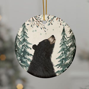 May include: A round ceramic ornament featuring a watercolour illustration of a black bear looking upwards. The bear is surrounded by green pine trees and light blue snowflakes against a white background. A gold string is attached for hanging.