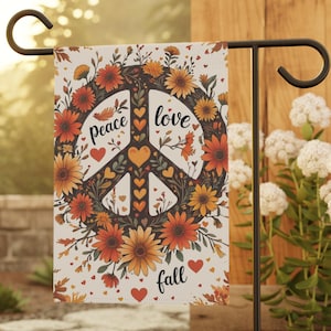 Pode incluir: Uma bandeira de jardim retangular com um símbolo da paz central rodeado por flores e folhas de outono em tons de laranja, amarelo e castanho. O texto "peace love fall" está impresso em preto, com pequenos corações vermelhos e laranjas.