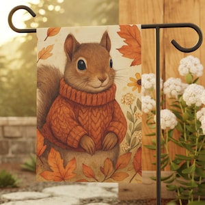 May include: A rectangular garden flag featuring an illustration of a brown squirrel wearing a textured orange cable-knit sweater. The squirrel is surrounded by orange and yellow autumn leaves and small yellow flowers on a cream-colored background.