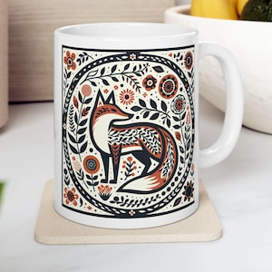 Peut inclure: Mug en céramique blanche avec un motif de renard stylisé dans un cadre floral circulaire. Le renard est représenté en orange, noir et crème, entouré de motifs floraux et de feuilles détaillés. La tasse a une anse blanche et repose sur un dessous de verre beige.
