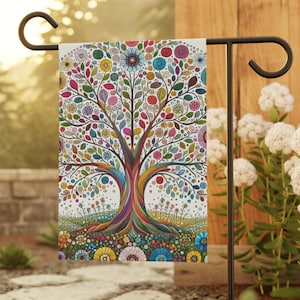 May include: A garden flag with a detailed illustration of a tree with colourful leaves and flowers. The tree's trunk and roots are multi-coloured, and the flag is displayed on a black metal stand. The design is set against a natural outdoor backdrop.
