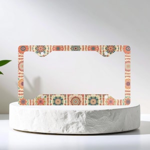 May include: A decorative license plate frame with a floral pattern in shades of orange, blue, and green on a cream background. The frame is displayed on a white pedestal against a white backdrop.