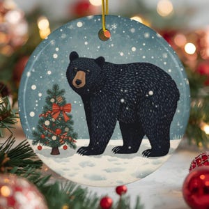 May include: A round ceramic ornament featuring a black bear standing in a snowy landscape next to a decorated Christmas tree with red ornaments and a red bow. The background is a soft blue with white snowflakes falling throughout the scene.