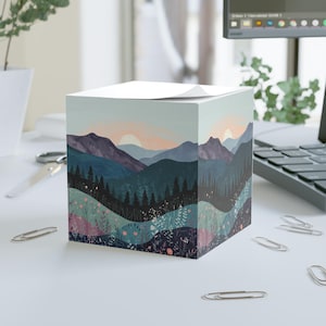 Peut inclure: Un bloc-notes cubique avec un paysage de montagnes, d'arbres et un coucher de soleil dans des tons de bleu, de violet et d'orange. Le bloc-notes est posé sur une surface blanche avec des trombones.