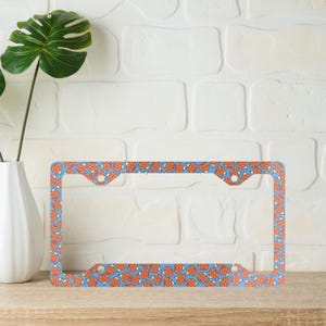 May include: A blue license plate frame with a repeating pattern of orange and white designs. The frame is rectangular with rounded corners and holes for mounting. The background is a light blue color.