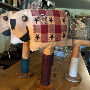 May include: Three whimsical fabric animal sculptures with bell accents and wooden spool legs. The central sculpture features a red and black checkered patch with black star accents and a circular tag with the number "1928". The sculptures are displayed on a wooden table.
