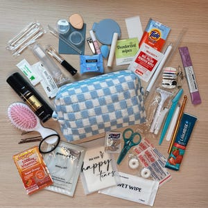 Puede incluir: Una colección de artículos de aseo y accesorios de viaje, que incluye un neceser a cuadros azules y blancos, un cepillo para el pelo, un cortaúñas y varios paquetes de artículos de cuidado personal. La imagen también incluye una pequeña botella de loción corporal y un paquete de Emergen-C.