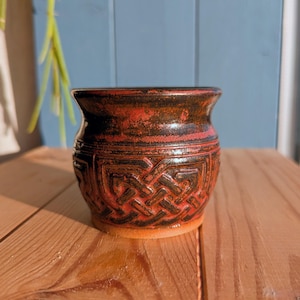 Puede incluir: Una maceta de cerámica hecha a mano con un esmalte marrón rojizo y un diseño detallado de nudo celta. La maceta tiene una boca ancha y un cuerpo redondeado, perfecta para guardar pequeñas plantas o artículos decorativos.
