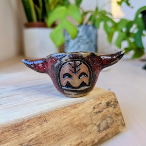 May include: A small, handmade ceramic bowl with a unique design. The bowl is mainly red with brown and black accents. It features a carved face design with a leaf in the center. The bowl has two horn-like extensions on either side.