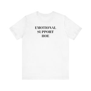Könnte beinhalten: Weißes T-Shirt mit dem schwarzen Text "EMOTIONAL SUPPORT HOE". Das T-Shirt hat kurze Ärmel und einen Rundhalsausschnitt. Der Text ist mittig auf der Vorderseite des Shirts platziert.