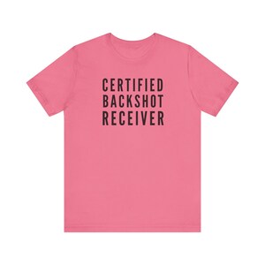 May include: A pink t-shirt with the text "CERTIFIED BACKSHOT RECEIVER" printed in black. The shirt has short sleeves and a crew neck. The text is centered on the front of the shirt.