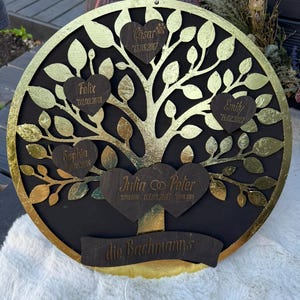 Könnte beinhalten: Runde Wanddekoration mit einem Familienstammbaum, mit schwarzem Hintergrund und goldfarbenem Baum und Blättern. Der Baum hat herzförmige Namensschilder mit Namen und Daten. Am unteren Rand befindet sich ein Banner mit der Aufschrift "die Bachmanns".
