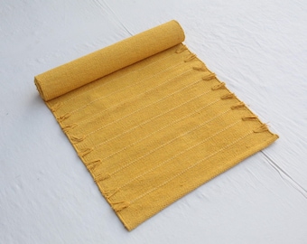May include: A rolled-up, mustard-yellow woven rug with a fringed edge. The rug is made of a textured material and has vertical stripes. The rug is partially unrolled, revealing its full width. The background is a white wooden surface.
