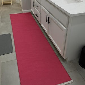 Op de afbeelding: Een lang, rechthoekig, fuchsia badkamertapijt met een gefranjerde rand. Het tapijt ligt voor een witte wastafel met zwarte hardware. De vloer is betegeld met neutraal grijs.