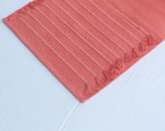 May include: Coral-colored rectangular rug with vertical ribbed texture and fringed edges. The rug has a woven design with subtle white stitching that accentuates the ribbed pattern. The tassels are at the short ends of the rug.