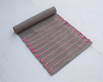 以下が含まれることがあります： 縦のピンク色のストライプと房が付いた、部分的に巻かれた灰色の織りラグ。ピンクのアクセントが見えます。白い表面にラグが置かれています。