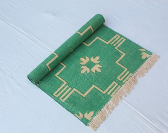 May include: A rolled-up green rug with a geometric design in a lighter tan color. The rug has a fringe on one end. The design includes a central floral-like pattern and a border of geometric shapes.