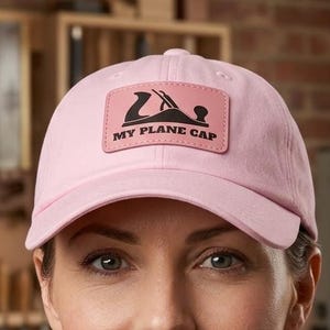 May include: A light pink baseball cap with a rectangular patch that reads "MY PLANE CAP" with a graphic of a plane. The cap is made of a soft material and has a curved brim. The background shows a workshop.