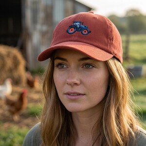 May include: A rust-colored corduroy baseball cap featuring an embroidered blue tractor with orange wheels. The cap has a curved brim and is worn by a person with long, light brown hair. The background includes a barn and a field.