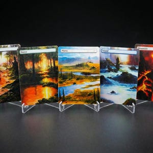 Peut inclure: Cinq cartes d'art paysagères, chacune présentée dans un présentoir en acrylique transparent. Les cartes représentent des scènes de marais, de forêt, de plaines, d'île et de montagne. L'art présente des couleurs vives et des images détaillées, avec des étiquettes de texte en haut de chaque carte.