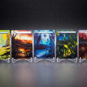Può includere: Cinque carte d'arte fantasy, ognuna raffigurante un paesaggio diverso: Foresta, Pianure, Isola, Palude e Montagna. Le carte sono esposte su supporti in acrilico trasparente su sfondo scuro. Ogni carta presenta colori vivaci e opere d'arte dettagliate.
